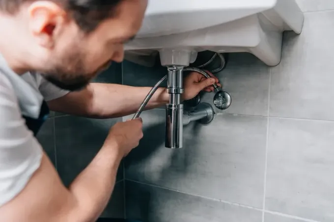 Persona arreglando tuberías bajo un fregadero blanco con una llave inglesa. Pared de azulejos grises.
