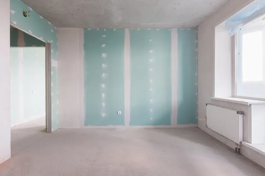 Habitación en construcción con paneles de yeso verde, puerta abierta y ventana con radiador.