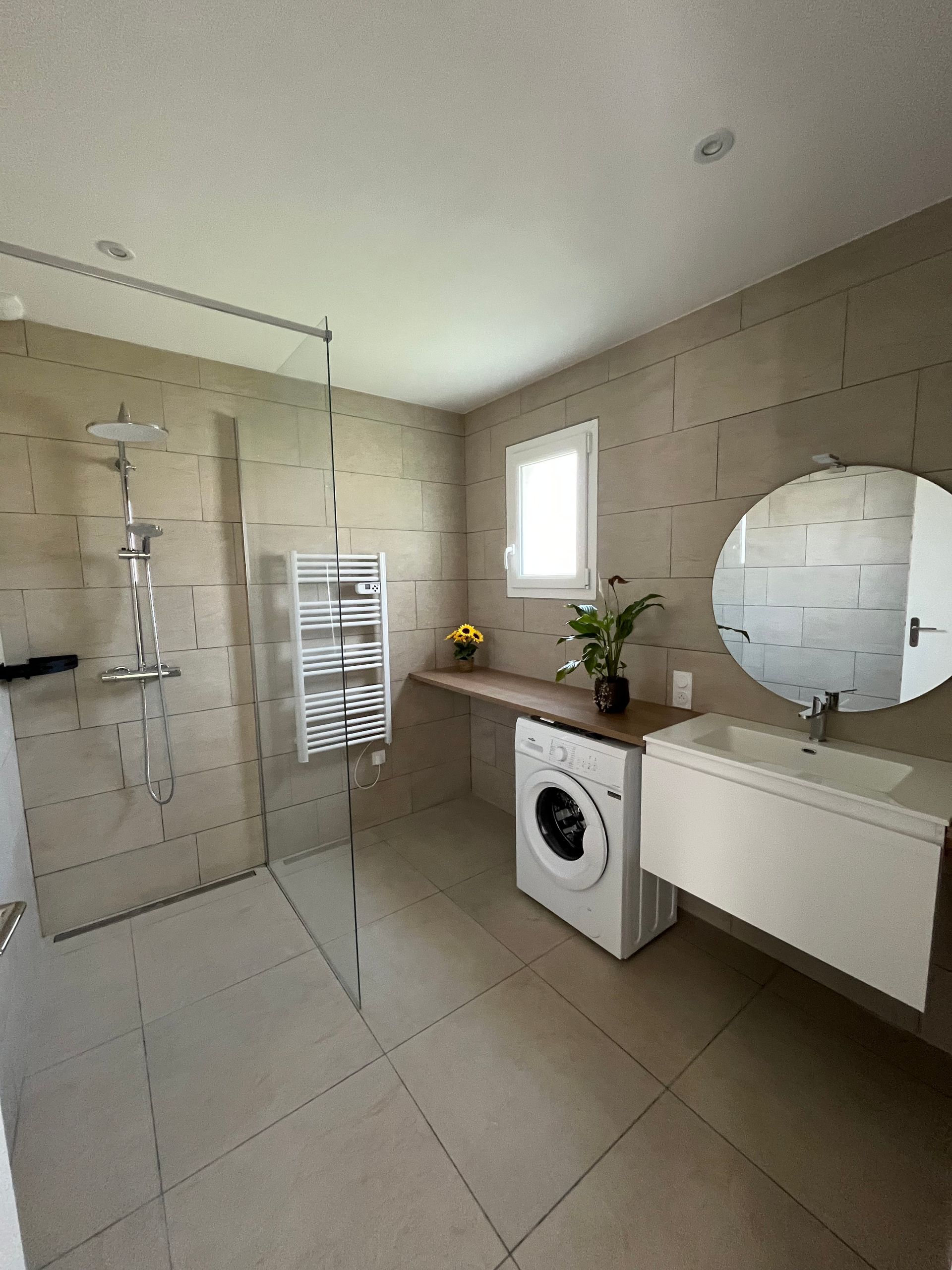 Salle de bain moderne avec douche, buanderie, lavabo et miroir rond. Tons neutres, fenêtre.