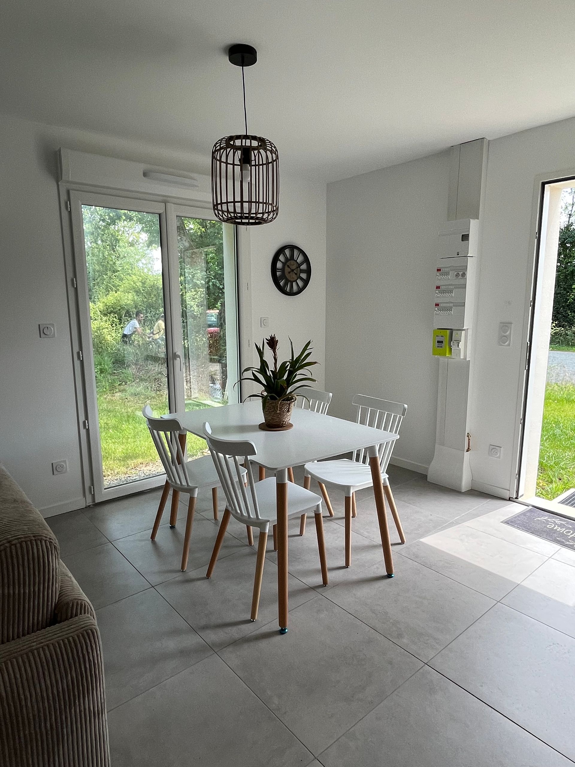 Salle à manger avec table blanche, chaises et suspension noire ; porte donnant sur un jardin.