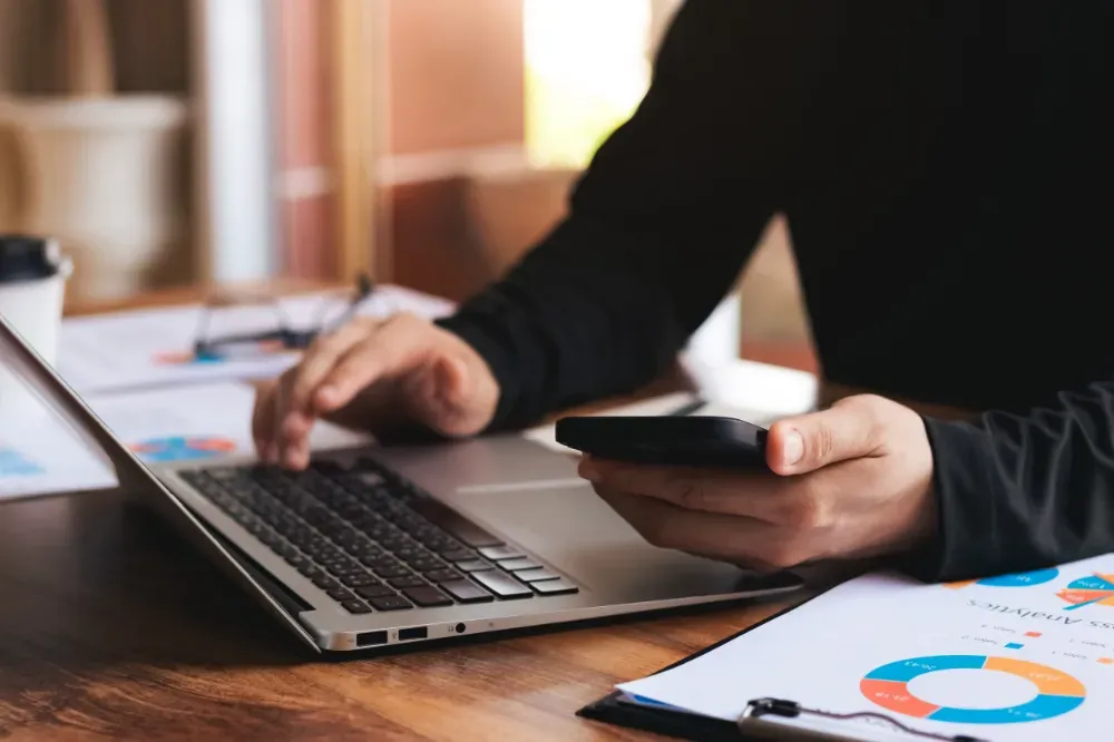 Persona trabajando en una computadora portátil y sosteniendo un teléfono, rodeada de gráficos y café.