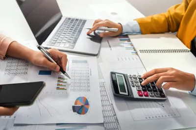 Manos trabajando con documentos, computadora portátil, calculadora y teléfono.