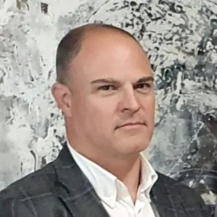 Hombre con blazer y camisa blanca, mirando al espectador. Fondo de arte abstracto gris y blanco.