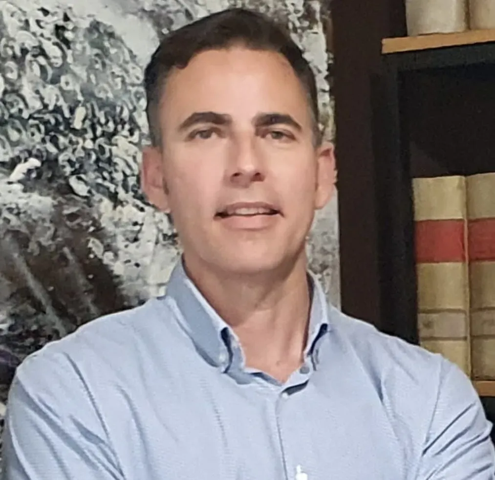 Un hombre con una camisa azul claro sonríe, en un espacio interior cerca de arte abstracto.