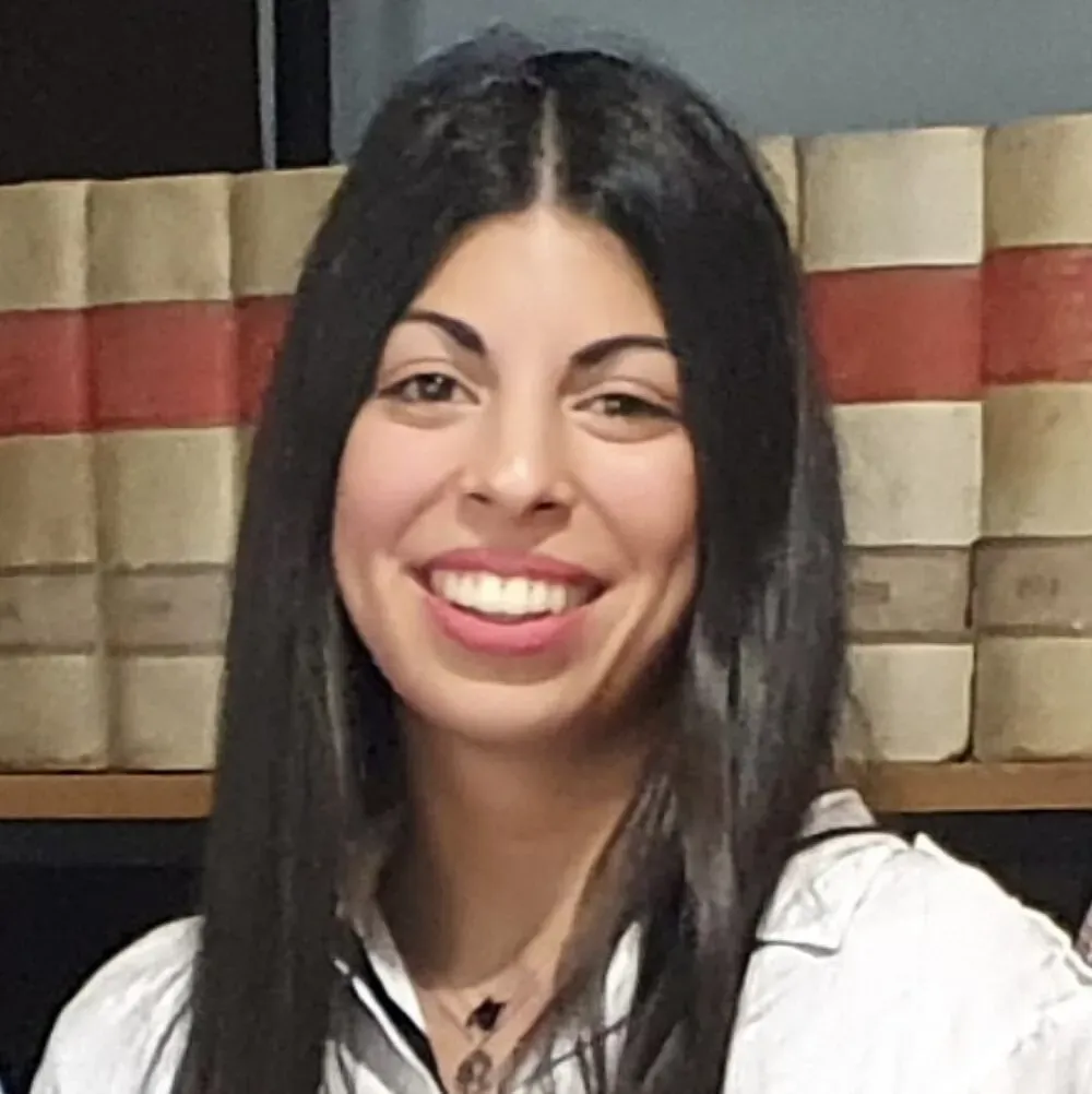 Una mujer con cabello largo y oscuro sonríe frente a una estantería, vistiendo una camisa con cuello.