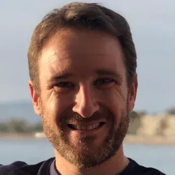 Un hombre con cabello castaño y barba sonríe ampliamente al aire libre cerca del agua.