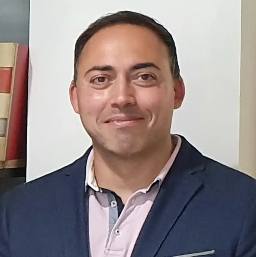 Hombre con blazer azul y camisa rosa sonríe, fondo neutro.