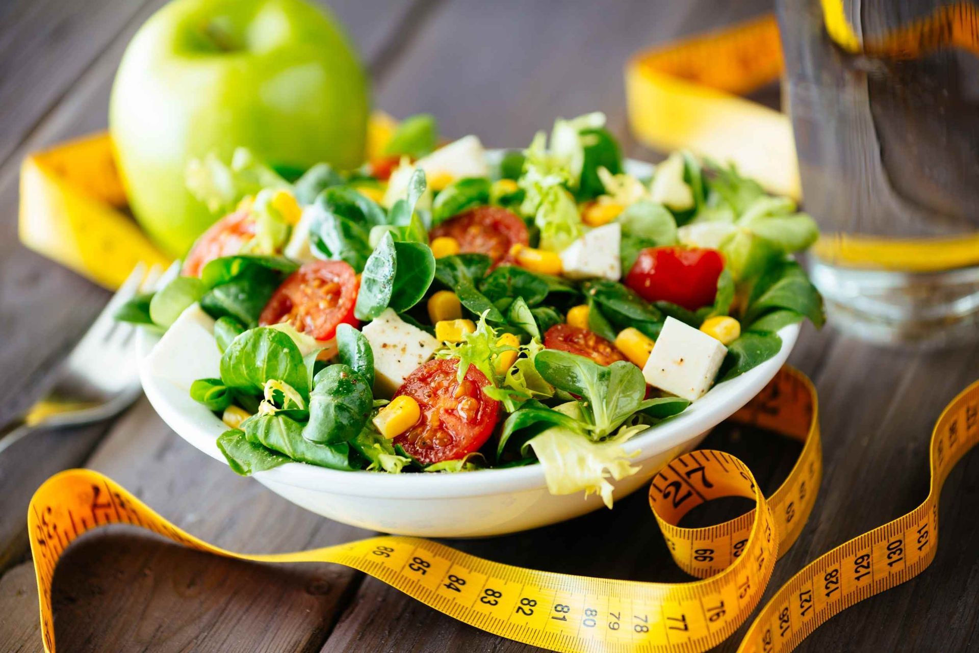 Un bol de ensalada junto a una cinta métrica y una manzana.