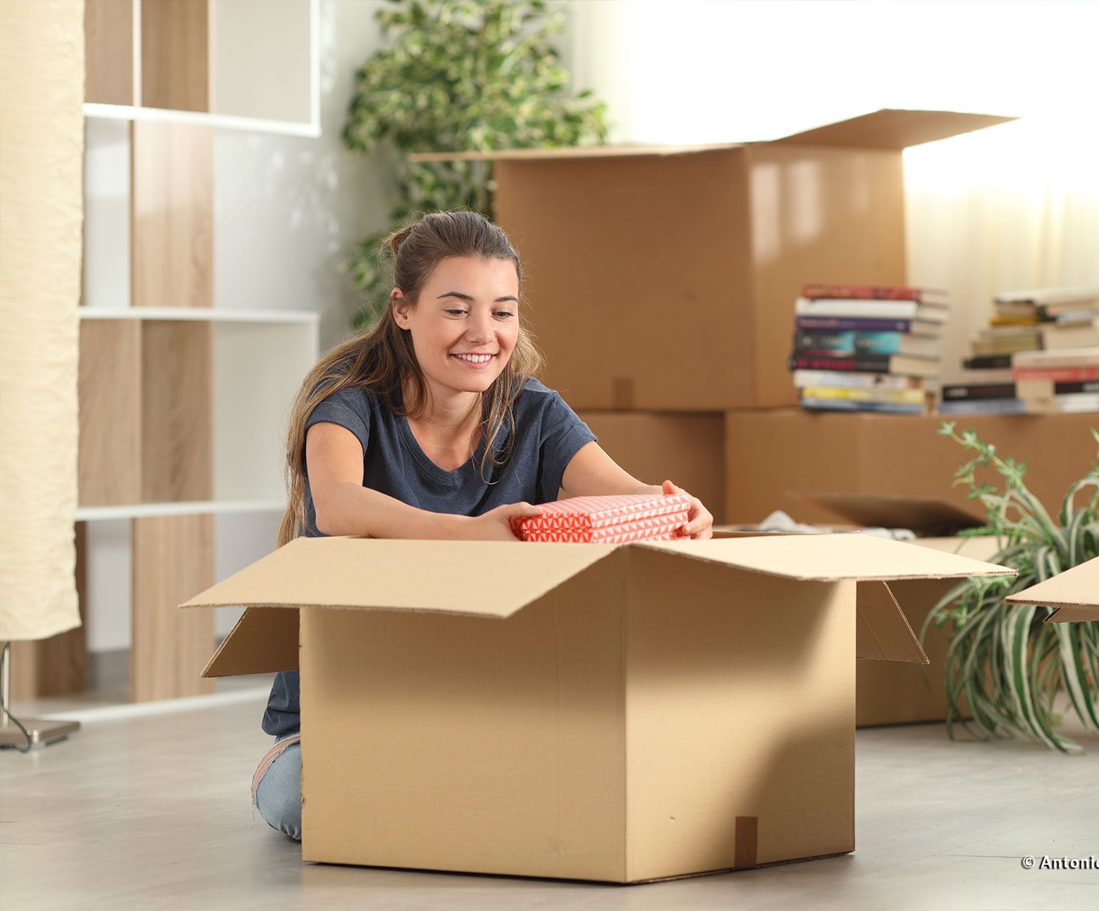 eine junge Frau packt einen Karton in ihrer ersten eigenen Wohnung aus und freut sich über den Inhalt