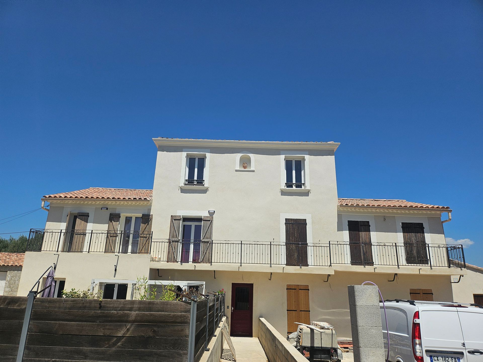 Maison blanche à deux étages avec toit de tuiles rouges, volets marrons et balcons sous un ciel bleu limpide.