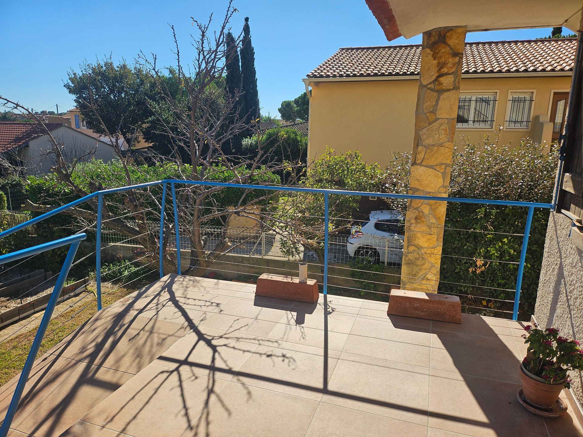 Terrasse avec balustrade bleue, supports en briques, donnant sur un jardin arboré avec un bâtiment.