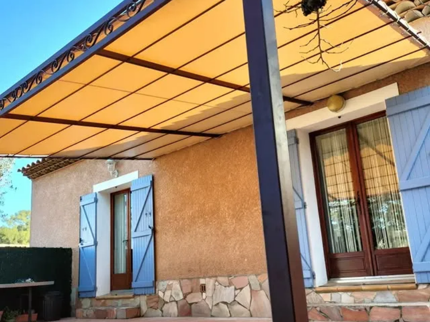 Un auvent beige recouvre une maison aux volets bleus et à la porte marron.