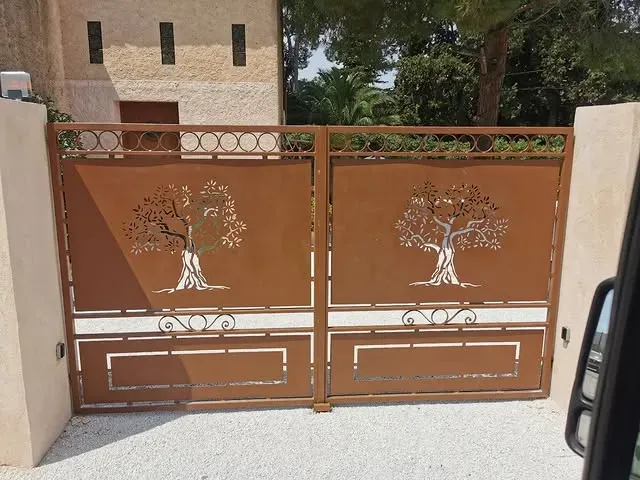 Portail en métal marron orné d'un motif d'arbre découpé au laser, encastré dans des murs en pierre beige et une allée de gravier.