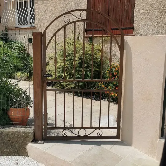 Portail en fer forgé marron à arc surbaissé, donnant sur un jardin.