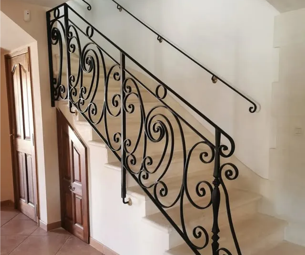 Escalier avec rampe en fer forgé noir ouvragée et mur blanc ; une porte en bois est visible.