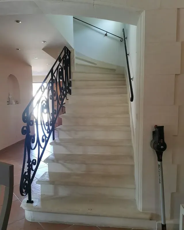 Escalier en marbre avec rampe en fer forgé ouvragée et main courante noire. Un aspirateur est appuyé contre le mur.