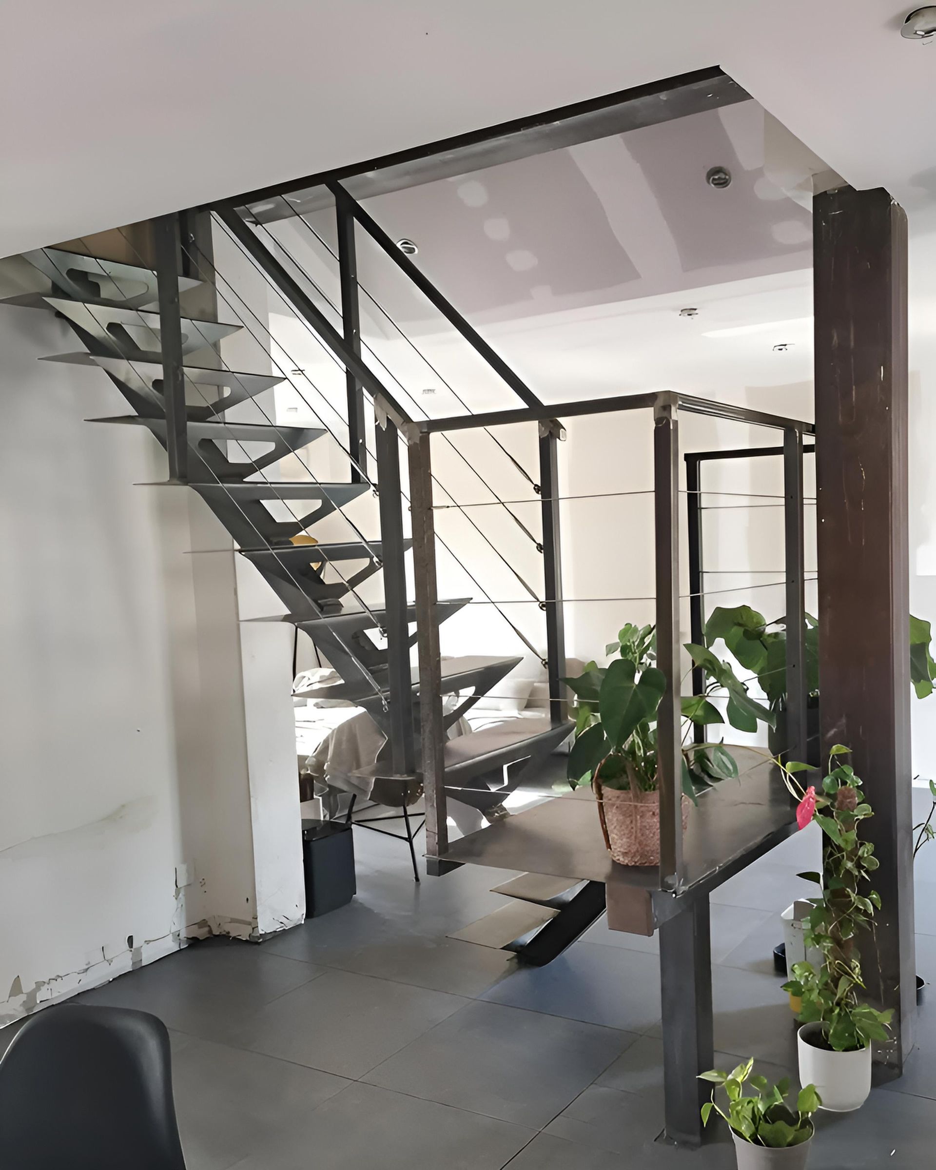 Escalier métallique menant à une plateforme avec des plantes en pot ; sol gris, murs blancs.