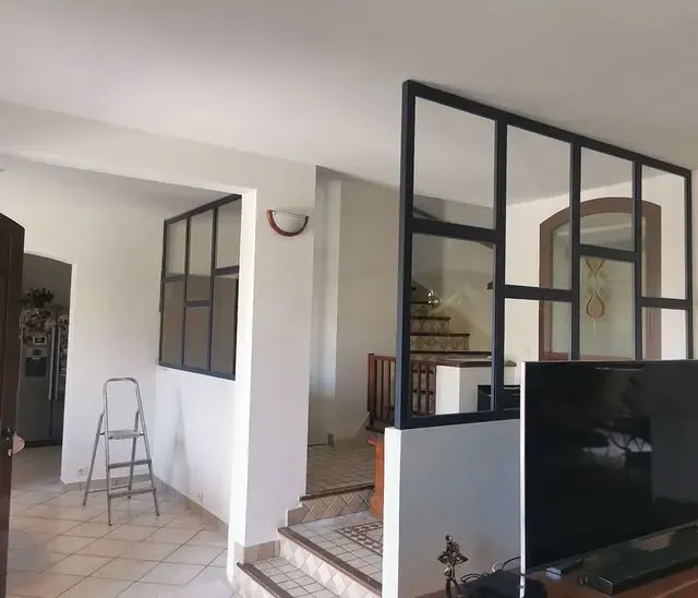 Vue intérieure d'une maison avec une verrière à cadre noir, un escalier et une grande télévision. Murs blancs et sols carrelés.