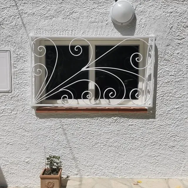 Fenêtre blanche ornée de ferronneries décoratives sur un mur blanc texturé. Une petite plante en pot est posée en dessous.