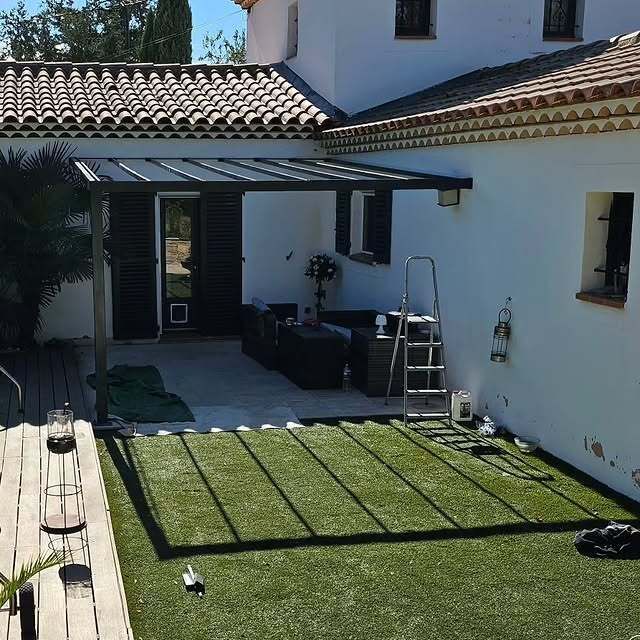 Une pergola ombrage une terrasse meublée de mobilier sombre. Du gazon synthétique recouvre un espace délimité. Bâtiment blanc, ensoleillé.