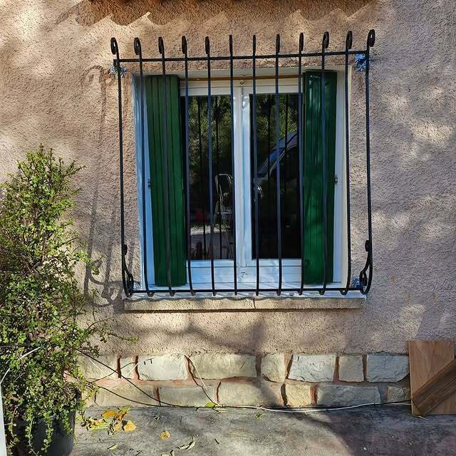Fenêtre à volets verts et barreaux de fer noirs. Un petit buisson vert se trouve à gauche.