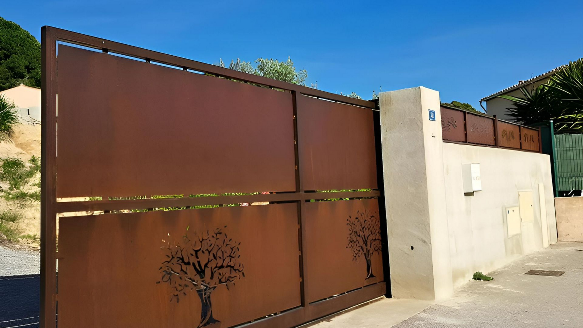 Un portail en métal marron orné de découpes d'arbres donne accès à une propriété, à côté d'un mur blanc.