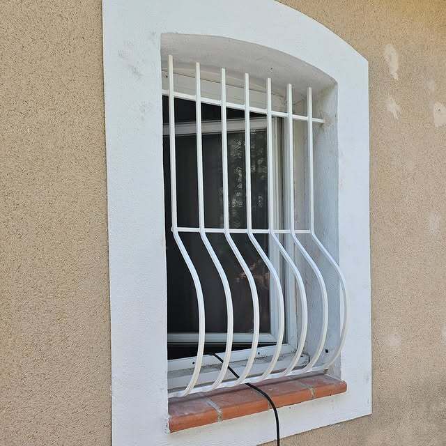 Fenêtre à barreaux blancs incurvés sur un bâtiment beige. Cadre cintré blanc avec appui en brique.