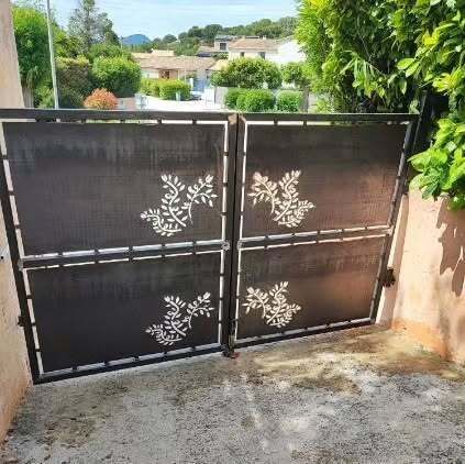 Portail en métal noir orné de découpes florales, fermé. Situé dans une allée avec des maisons en arrière-plan.