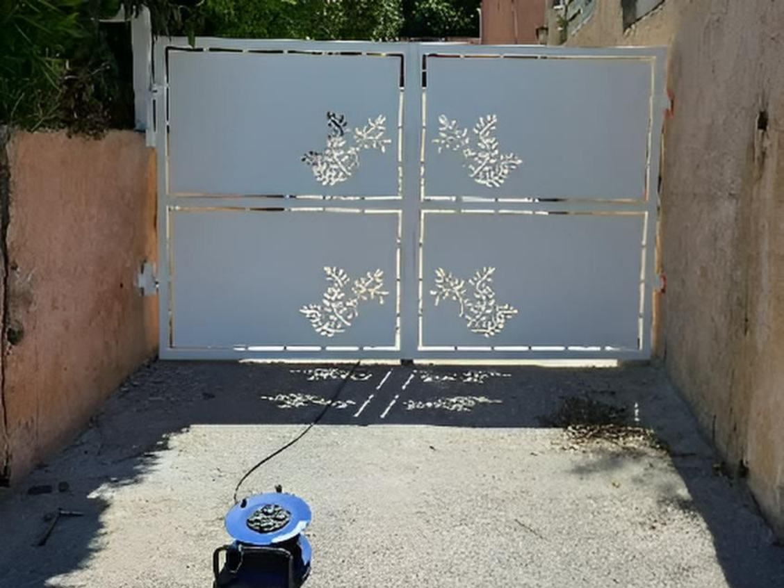 Portail en métal blanc orné de découpes florales, fermé, devant une allée. Ombres projetées sur le sol.