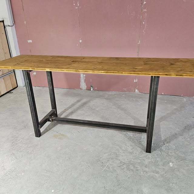 Table à plateau en bois et pieds en métal noir. Elle se trouve dans une pièce vide aux murs roses et au sol gris.