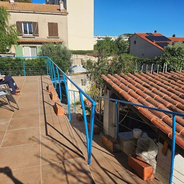 Terrasse sur le toit avec rambarde bleue, carrelage en terre cuite et bâtiments environnants sous un ciel bleu azur.