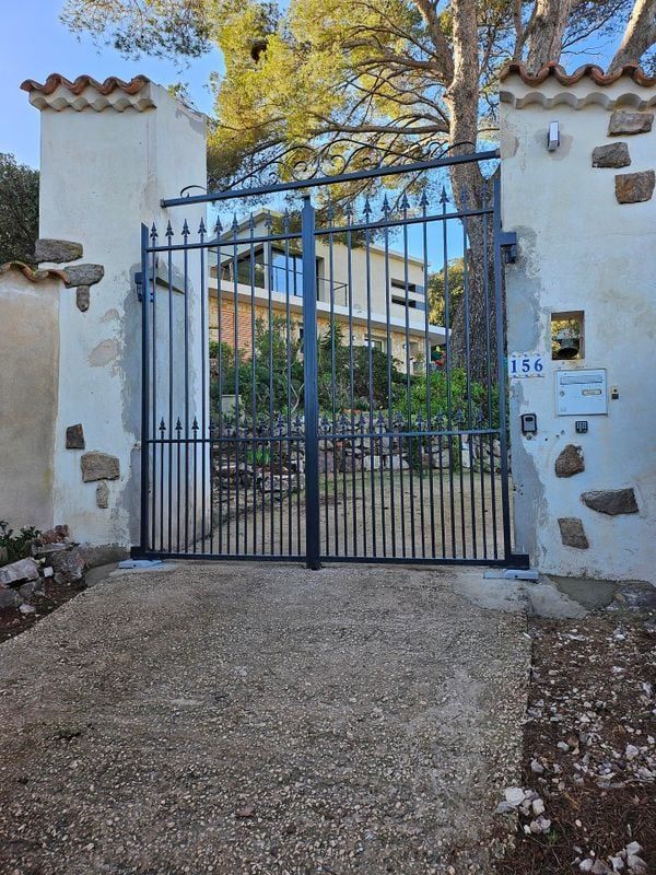 Un portail en métal noir à barreaux verticaux, placé entre des murs en pierre, donne accès à une maison.