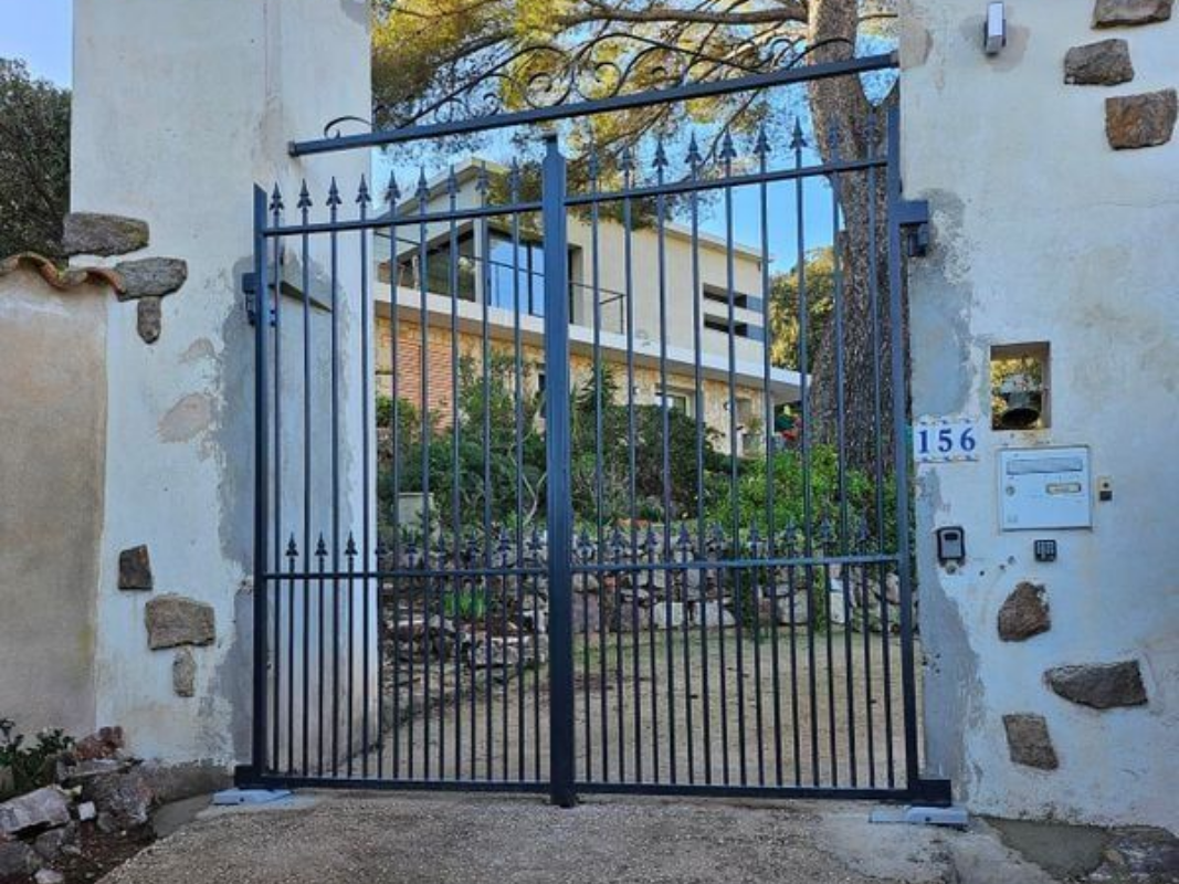 Un portail en métal noir marque l'entrée d'une maison au design moderne avec jardin. Le portail est encadré par des murs en pierre.