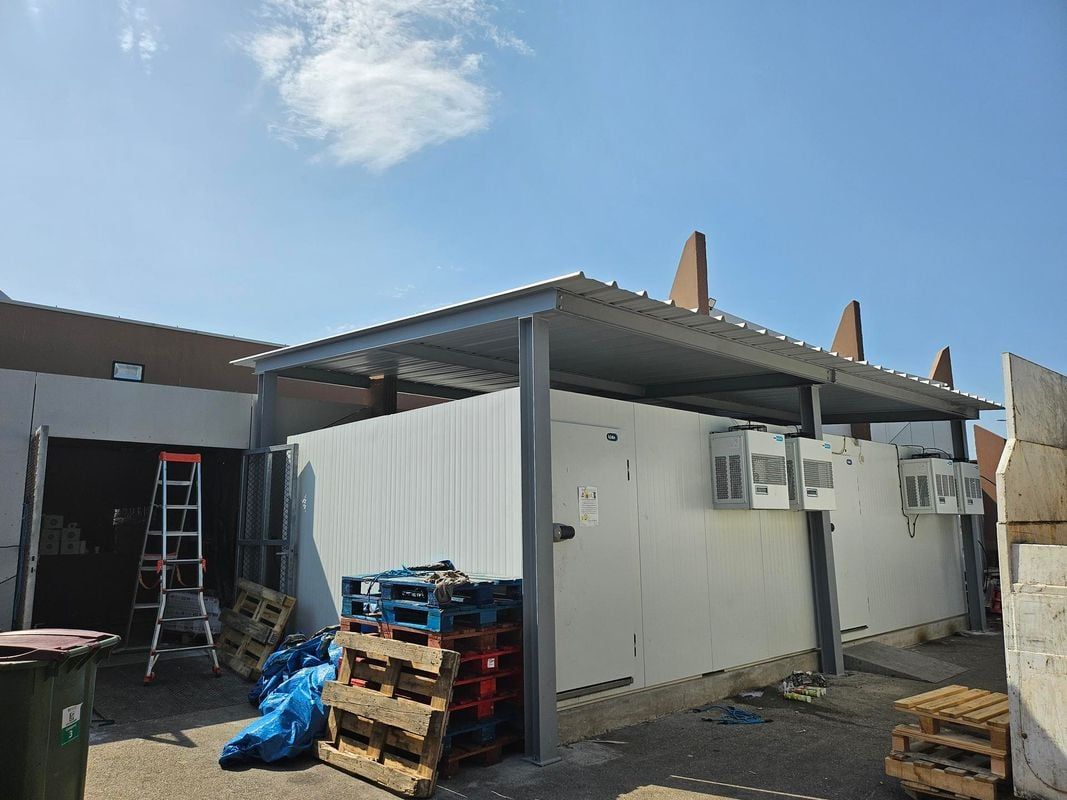 Vue extérieure d'un bâtiment avec un auvent métallique, des palettes et des poubelles.