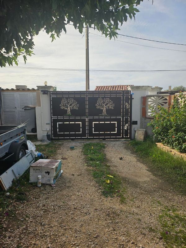 Portail en métal noir orné de motifs d'arbres, dans une allée de gravier, avec un poteau électrique et de la végétation environnante.