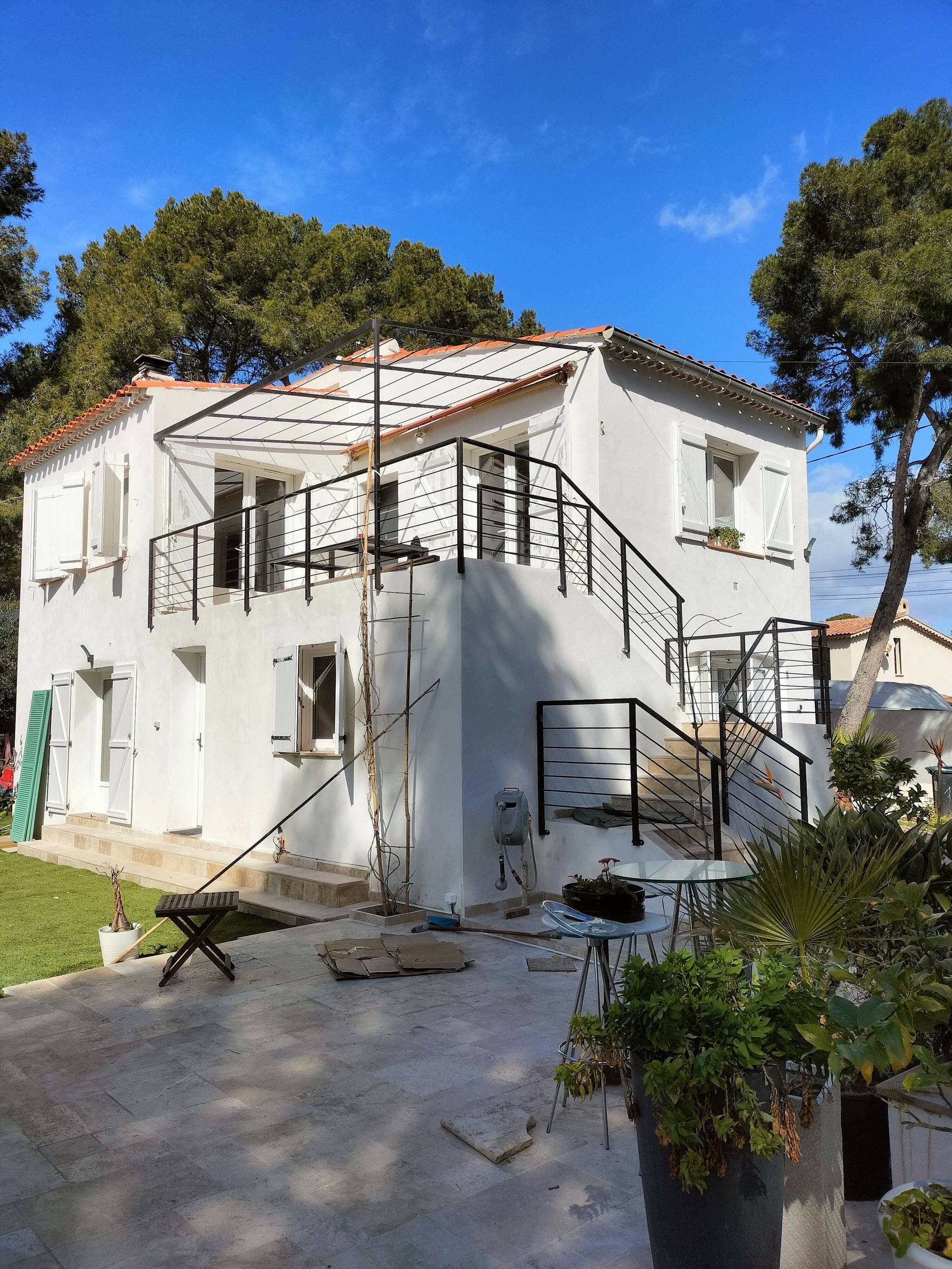 Maison blanche à deux étages avec balustrades et volets noirs, un escalier et des arbres environnants sous un ciel bleu.