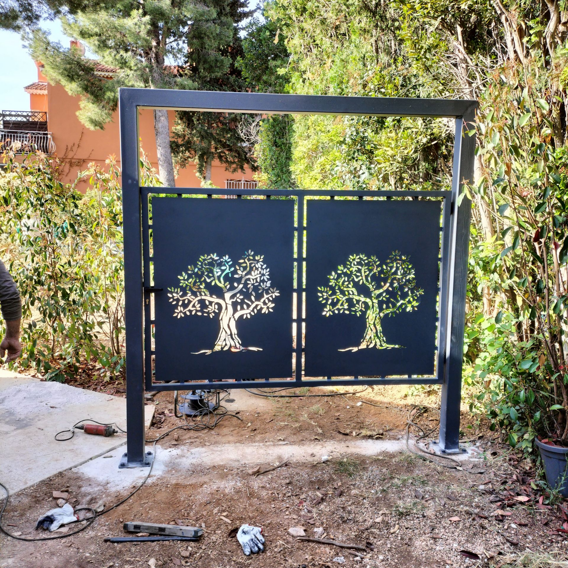 Portail en métal noir orné de silhouettes d'arbres devant un jardin.