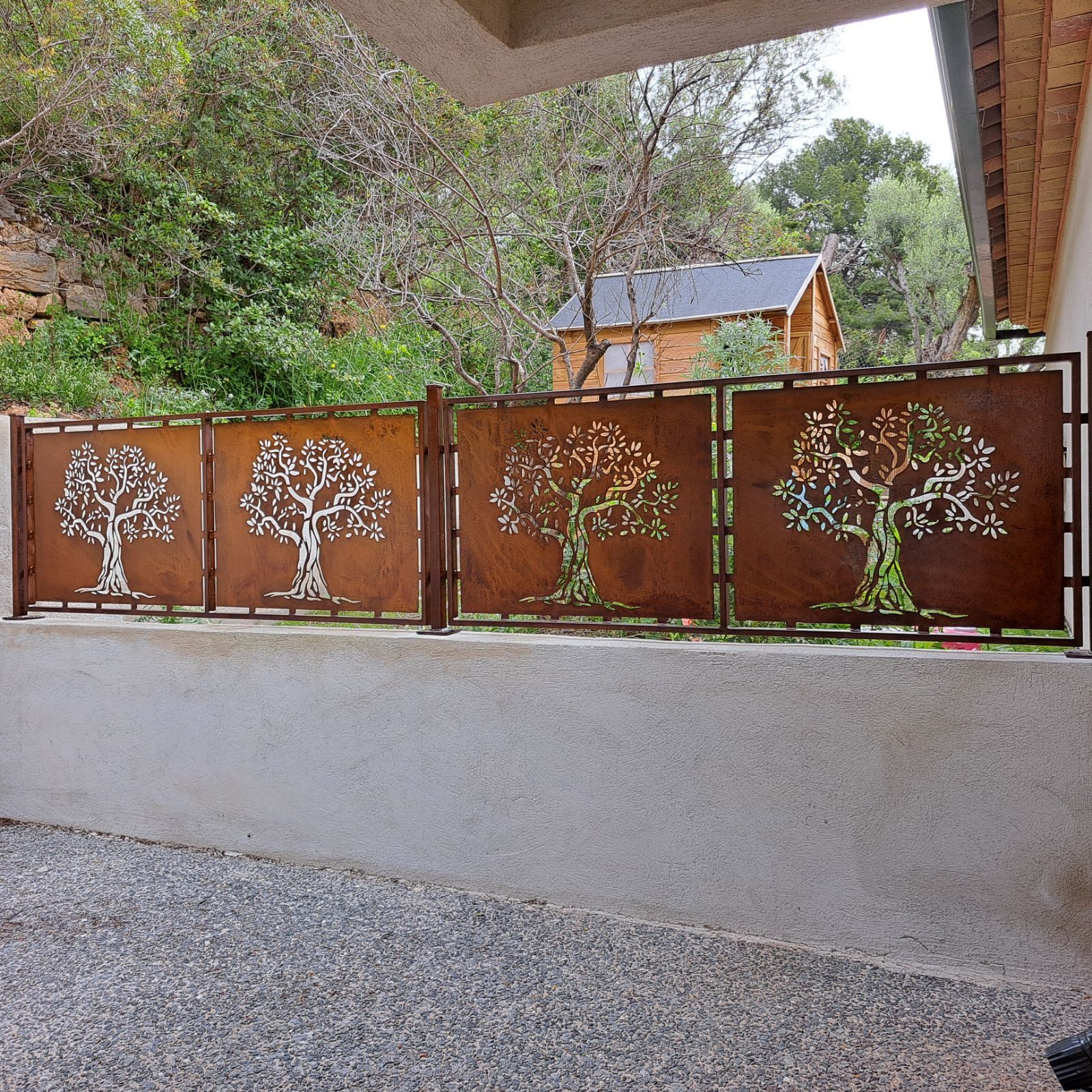 Quatre panneaux de clôture en métal couleur rouille ornés de motifs d'arbres, se détachant sur un fond boisé verdoyant.