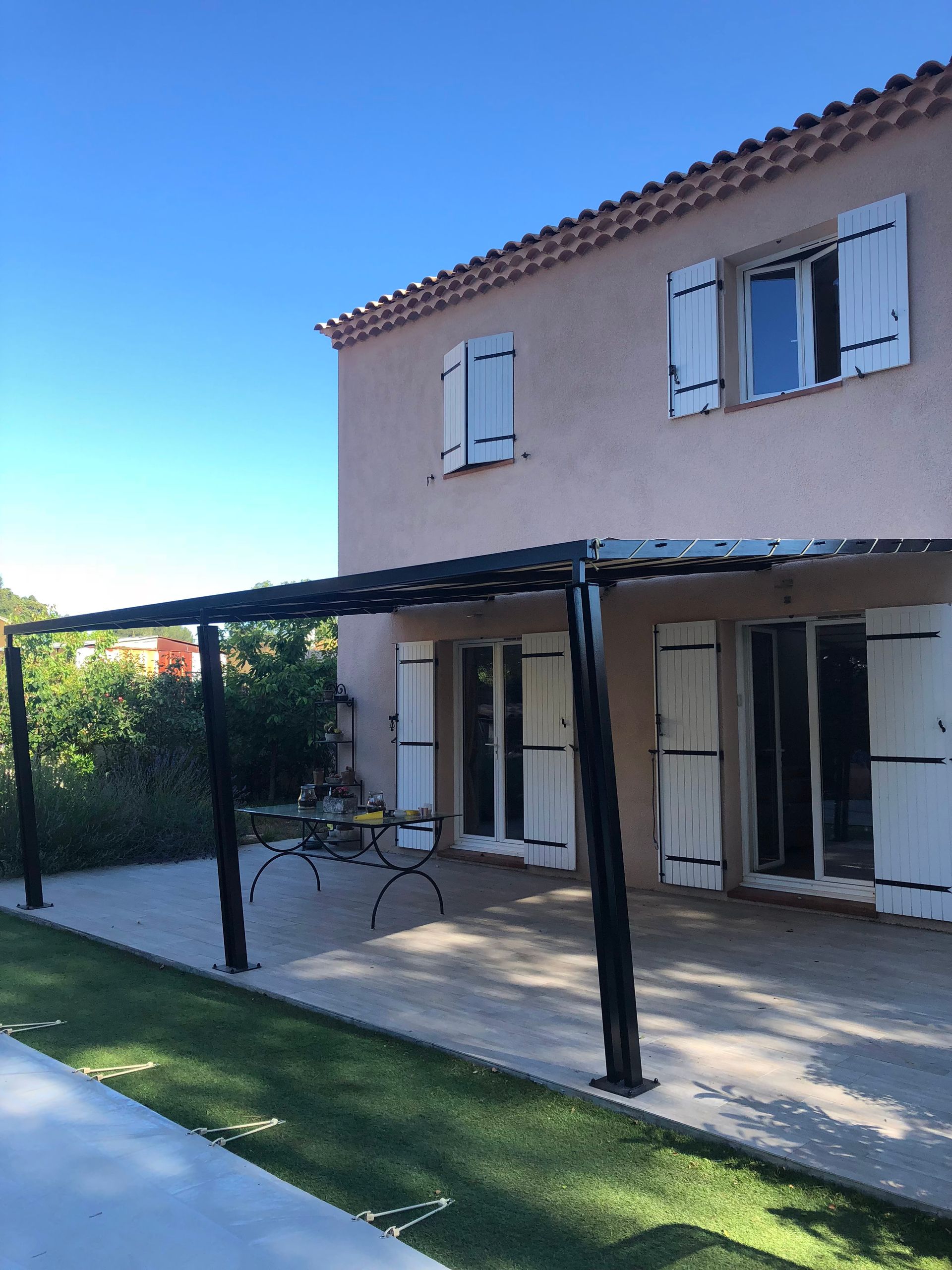 Une pergola noire s'étend depuis une maison rose aux volets blancs. Pelouse verte au premier plan, ciel bleu.
