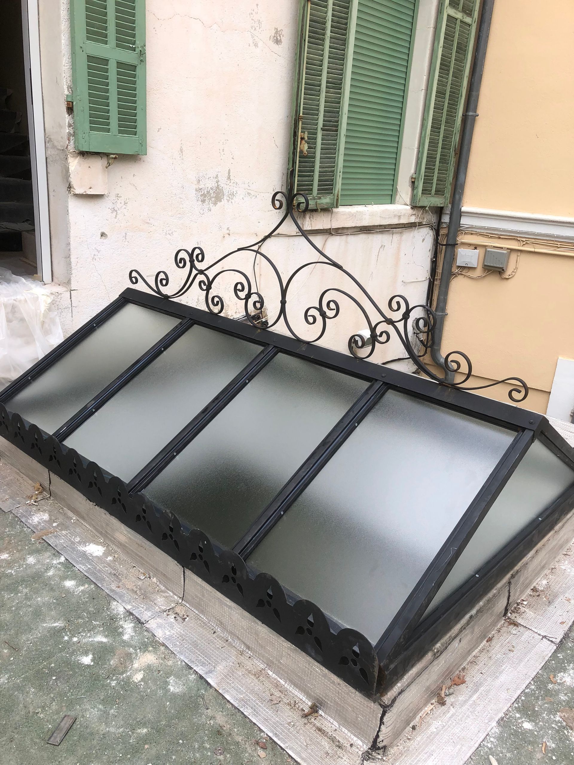 Puits de lumière orné de ferronneries noires, installé sur une surface en béton. Des volets verts sont visibles en arrière-plan.