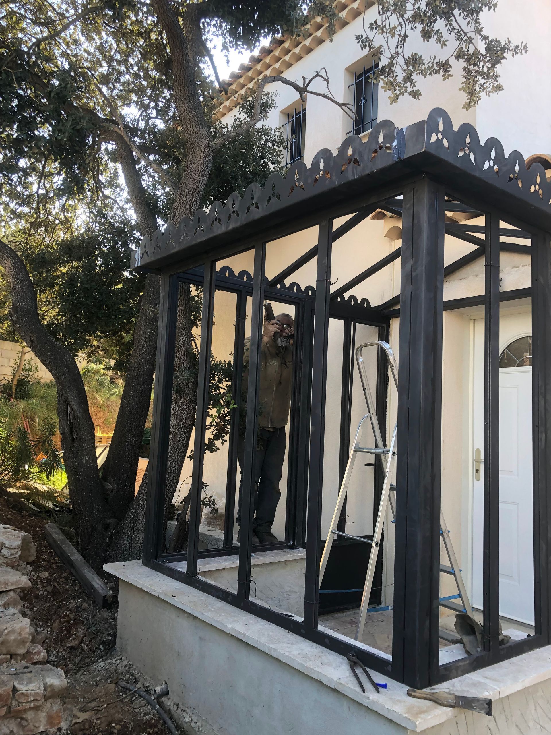 Un homme travaille sur une structure métallique noire fixée à un bâtiment blanc ; une échelle se trouve à l'intérieur, un arbre est situé à proximité.