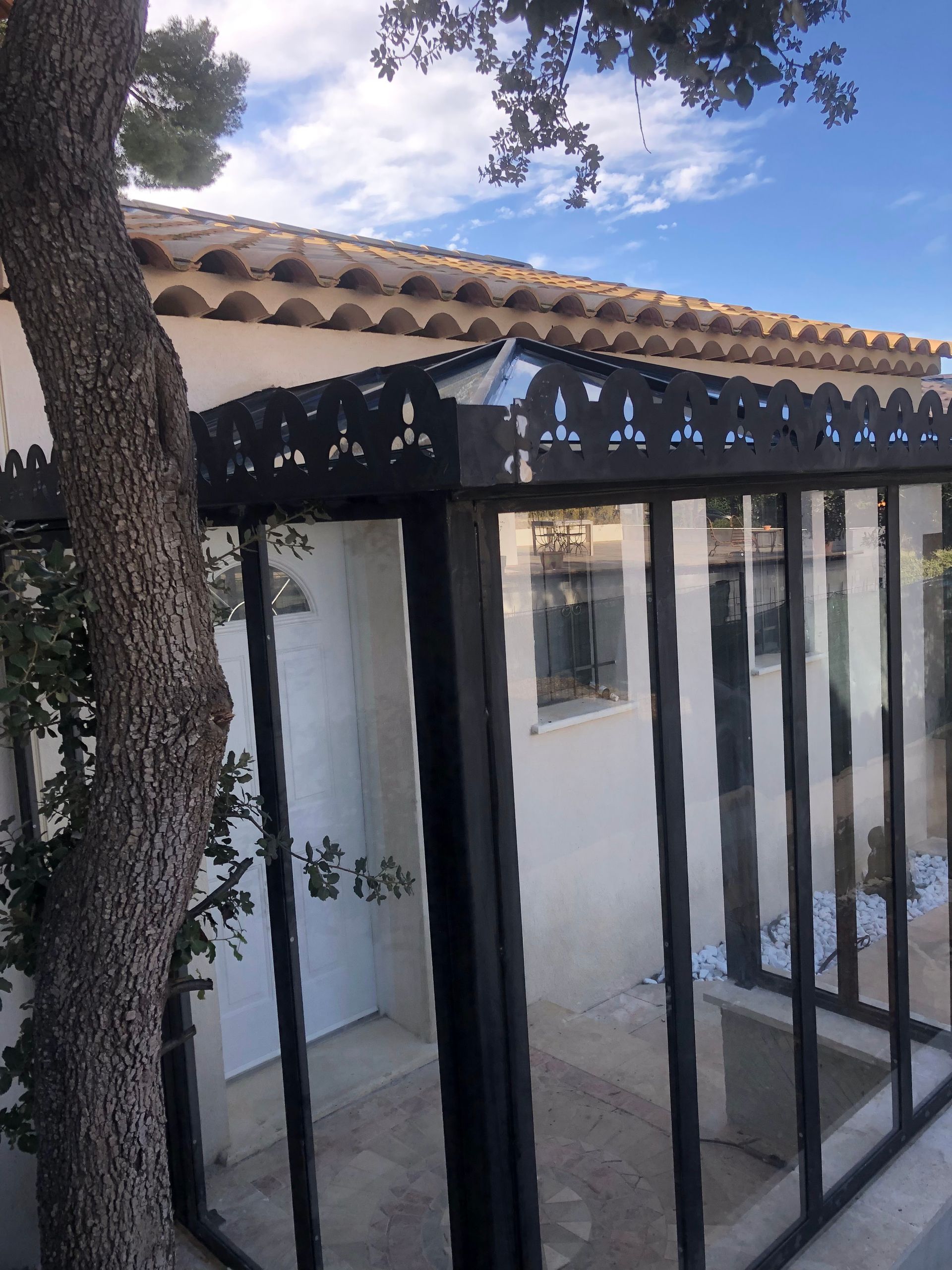 Enclos vitré avec structure en métal noir et détails de toiture ornementés, à côté d'un arbre.