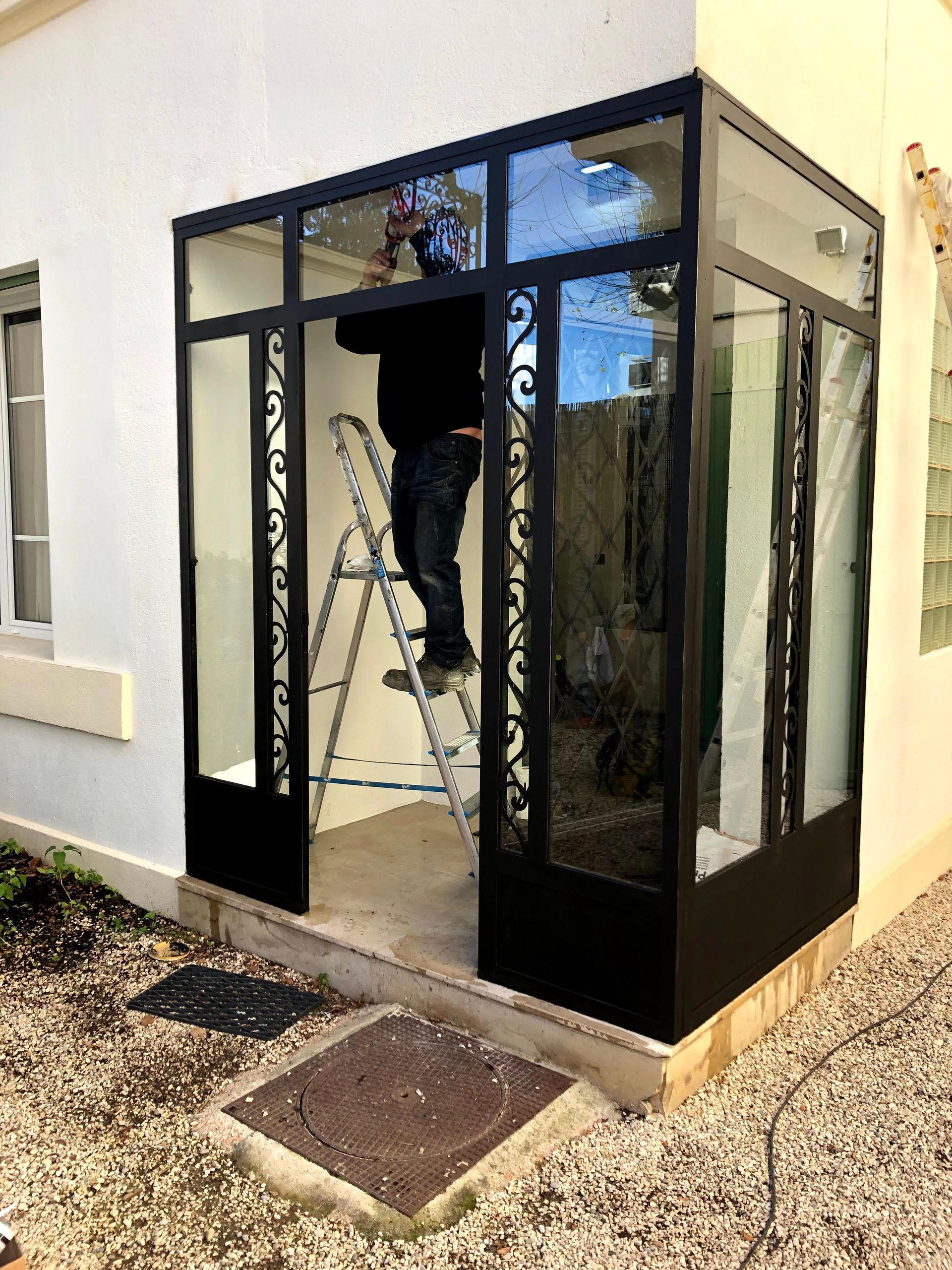 Une personne installe des panneaux de verre dans une entrée à cadre métallique noir. Façade du bâtiment blanche, échelle et découpes décoratives.