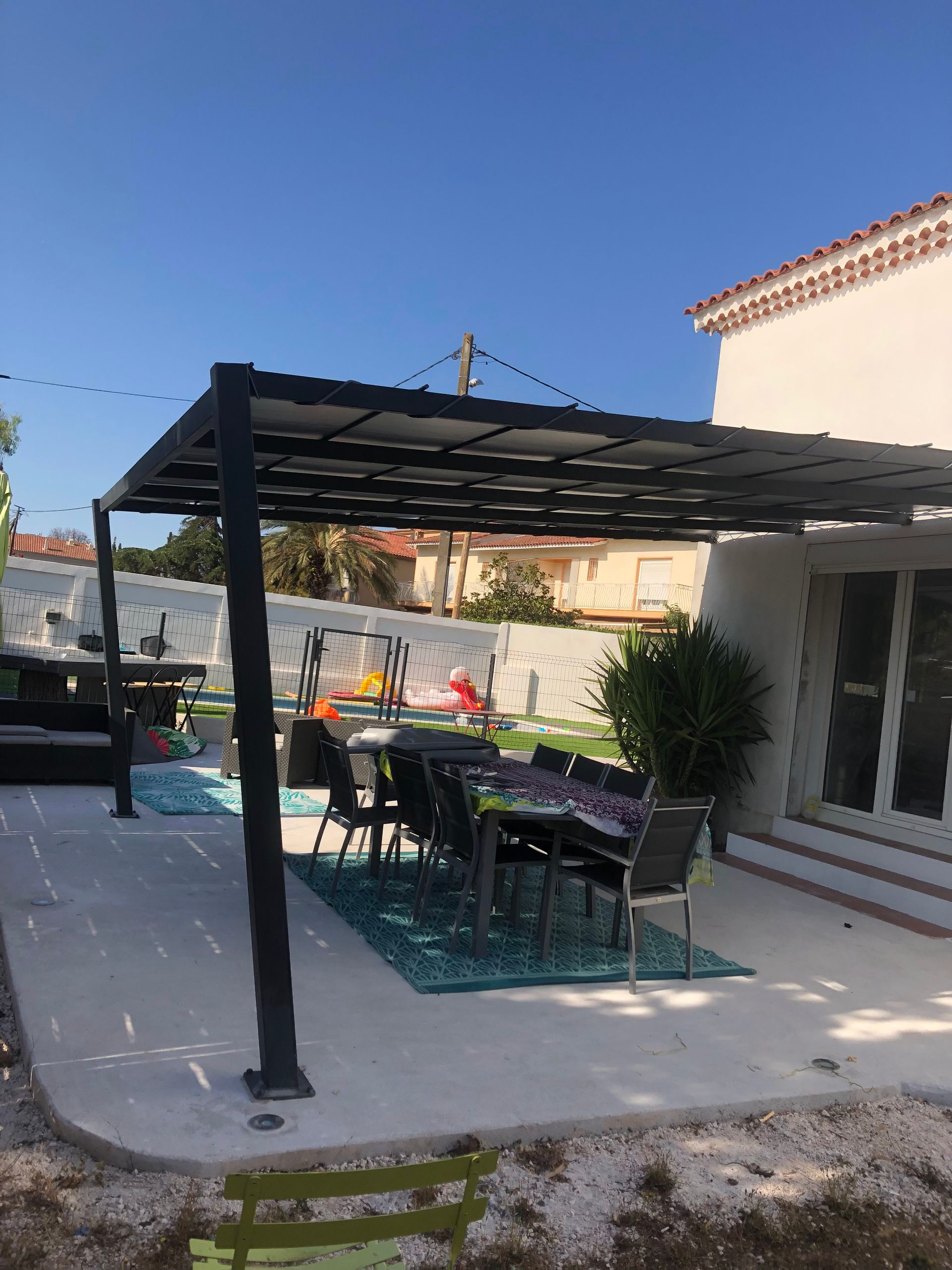 Pergola au-dessus d'une table et de chaises de jardin sur une terrasse en béton, avec une maison en arrière-plan.