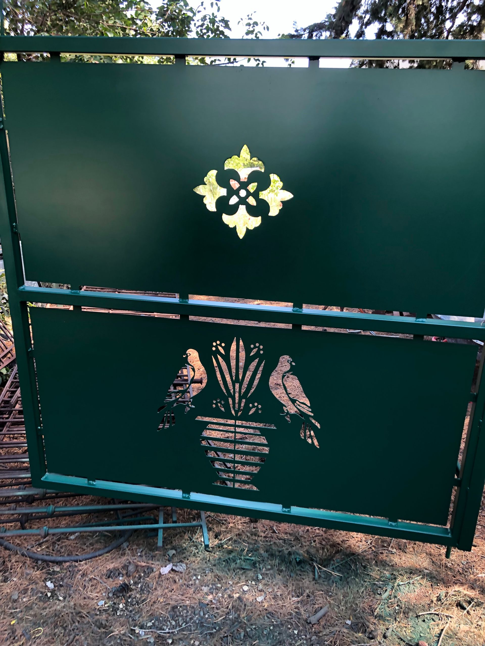 Portail en métal vert orné de découpes décoratives représentant une fleur de lys et un vase avec des oiseaux.