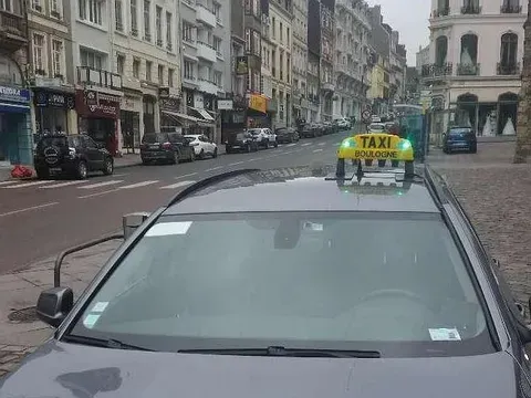 Un taxi avec une enseigne lumineuse sur le toit, dans une rue de la ville. Des immeubles bordent la route et des voitures sont garées à proximité.