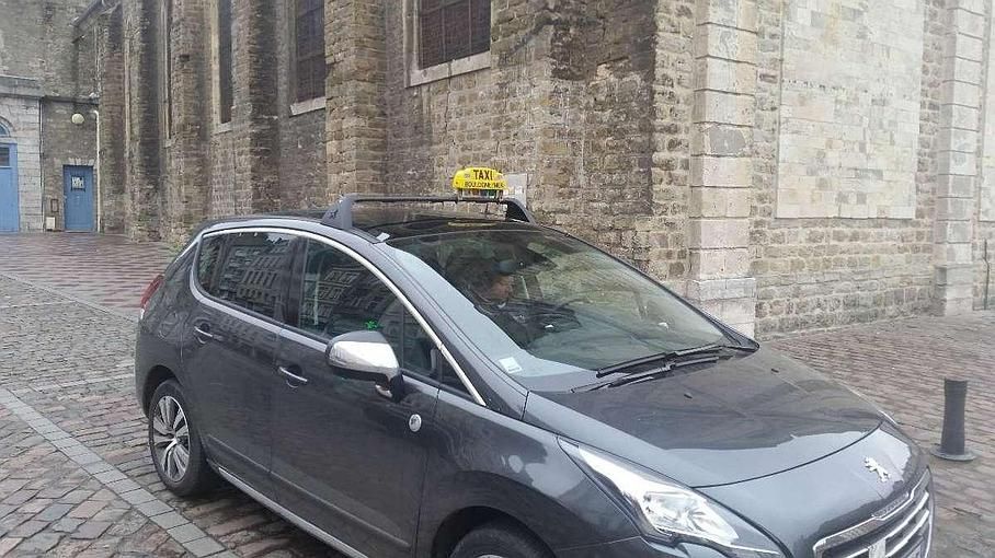 Un taxi gris est stationné dans une rue pavée, devant un immeuble en pierre. Il porte un panneau jaune sur le toit.