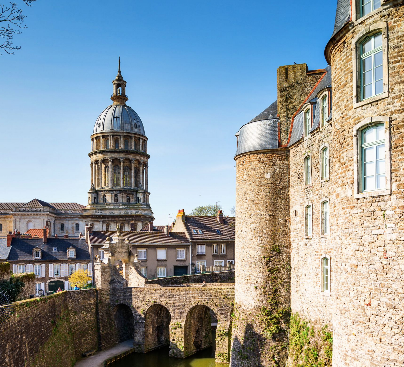 Château en pierre avec un bâtiment à dôme en arrière-plan, pont enjambant un cours d'eau, ciel bleu.