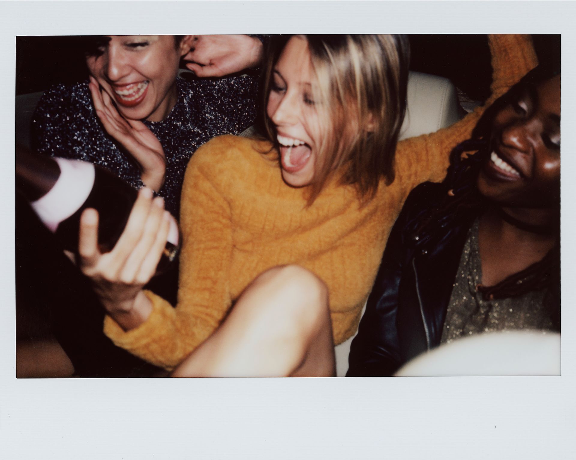 Trois femmes rient et célèbrent avec une bouteille de champagne dans une ambiance tamisée.