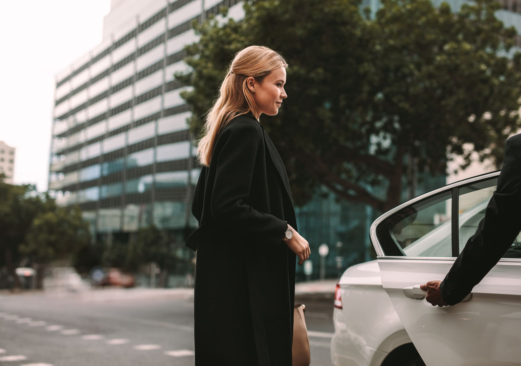 Une femme sort d'une voiture blanche, aidée par une personne qui lui ouvre la portière. Arrière-plan urbain.
